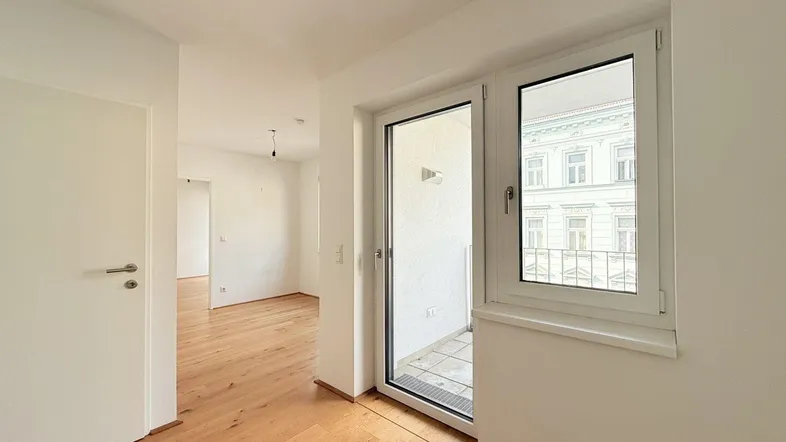 Helles Zimmer mit Holzboden und Zugang zu einem kleinen Balkon, bietet Blick auf die Stadt.