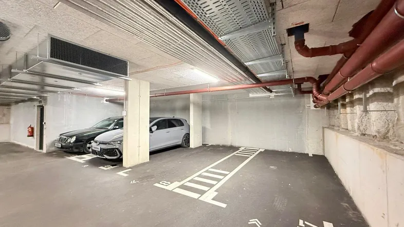 Tiefgarage mit mehreren Stellplätzen, sichtbaren Installationen an der Decke und Betonwänden.