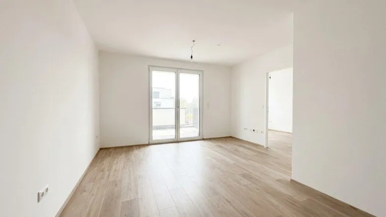 Helles, leeres Zimmer mit Holzboden und Zugang zu einem Balkon in einem Neubau.