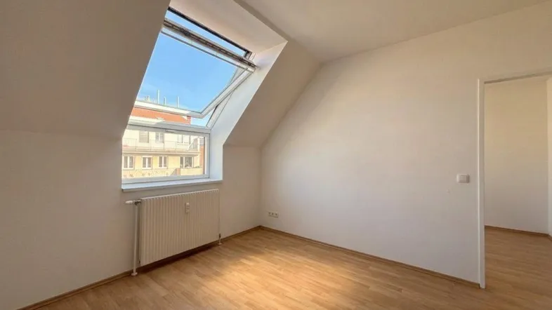 Helles Zimmer im Dachgeschoss mit Holzboden, Dachfenster und Blick auf umliegende Gebäude.