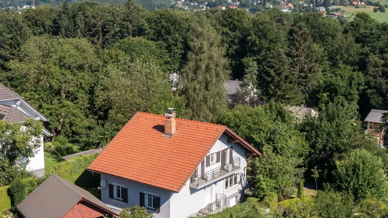 Luftaufnahme eines Einfamilienhauses mit rotem Ziegeldach und Carport, umgeben von viel Grün.