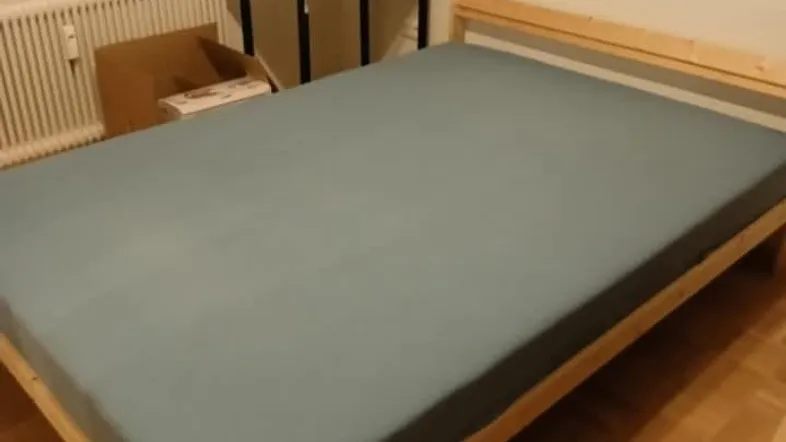 Helles Schlafzimmer mit Holzbettgestell, grauer Bettwäsche und Parkettboden.