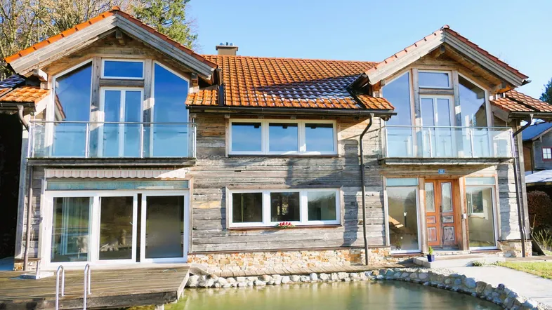 Rustikales Holzhaus mit großen Fenstern, Balkonen und einem kleinen Teich im Vordergrund.