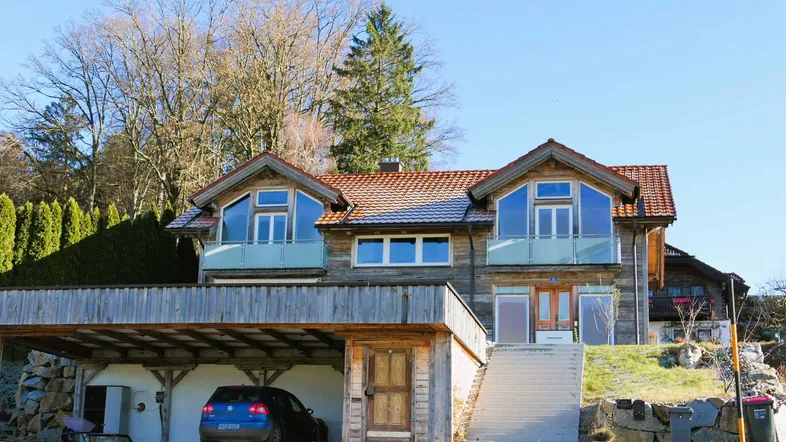 Modernes Holzhaus mit großen Fenstern, Balkonen und einem Carport, umgeben von Natur und Bäumen.