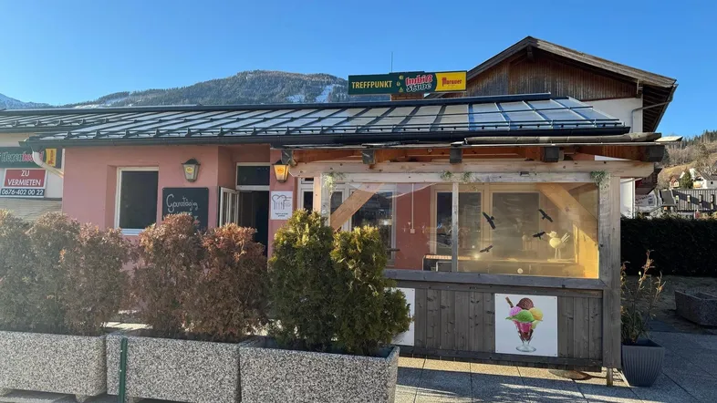 Außenansicht eines Imbisslokals mit überdachter Terrasse und Werbeschildern unter blauem Himmel.