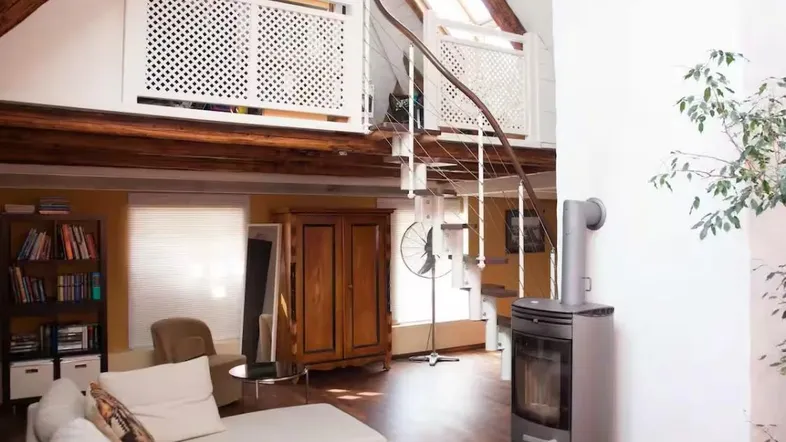 Helles Wohnzimmer mit hohen Decken, sichtbaren Holzbalken, Wendeltreppe und gemütlichem Holzofen.