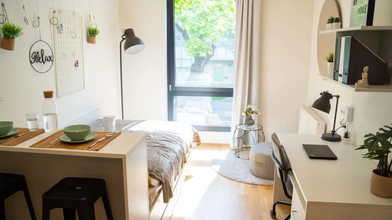 Helles Studio-Apartment mit Schlafbereich, Schreibtisch und Essgelegenheit am Fenster.