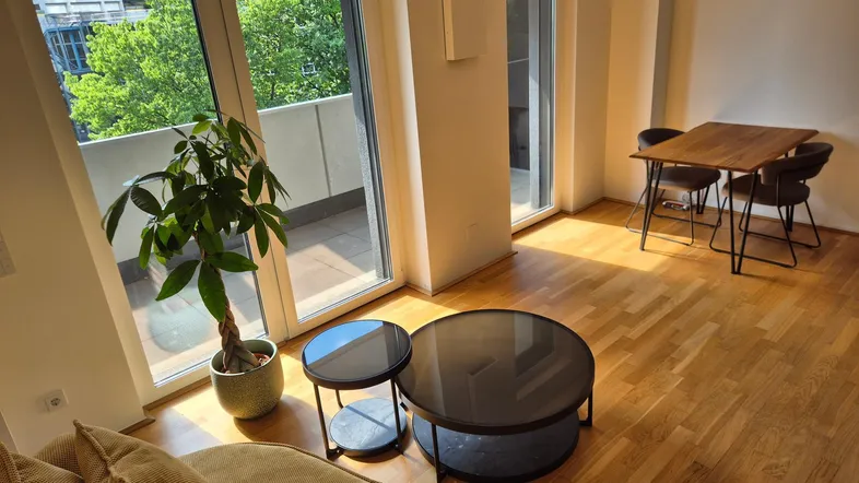 Helles Wohn- und Esszimmer mit Holzboden, Zugang zum Balkon und Blick ins Grüne.