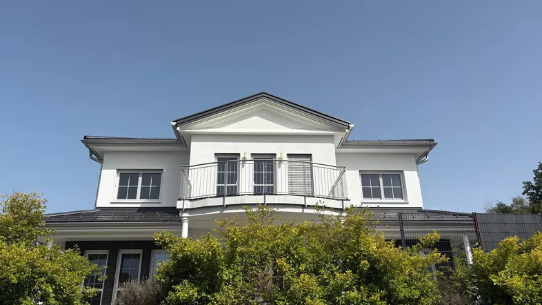 Modernes weißes Haus mit Balkon und dunklem Satteldach unter klarem blauen Himmel, umgeben von grünen Büschen.