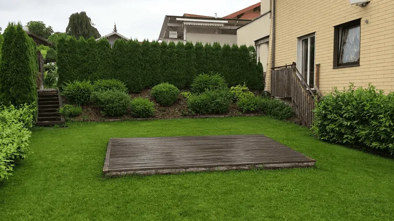 Gepflegter Gartenbereich mit Rasenfläche, Holzterrasse und grünen Hecken hinter dem Haus.