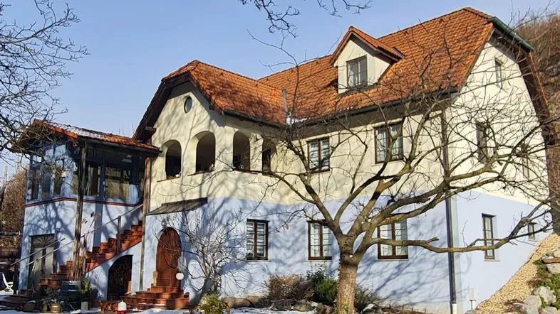 Traditionelles Haus mit hellblauer Fassade, rotem Ziegeldach und verschneitem Vorgarten unter blauem Himmel.