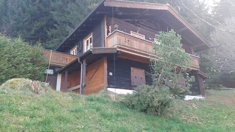 Traditionelles Holzchalet in alpiner Umgebung mit bewaldetem Hang im Hintergrund.