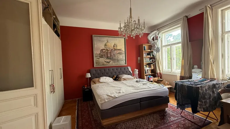 Geräumiges Schlafzimmer mit roten Wänden, Kronleuchter und Parkettboden, hell durch große Fenster.