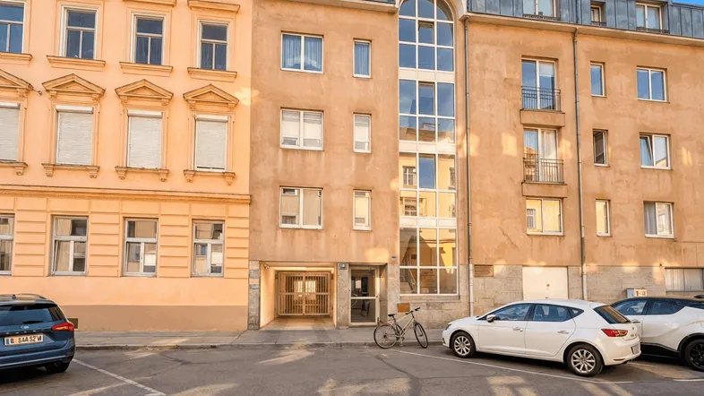 Fassade eines Mehrfamilienhauses mit angrenzender Straße und parkenden Autos bei sonnigem Wetter.
