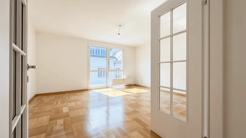 Helles, leeres Zimmer mit Fischgrätparkett und Zugang zu einem französischen Balkon.