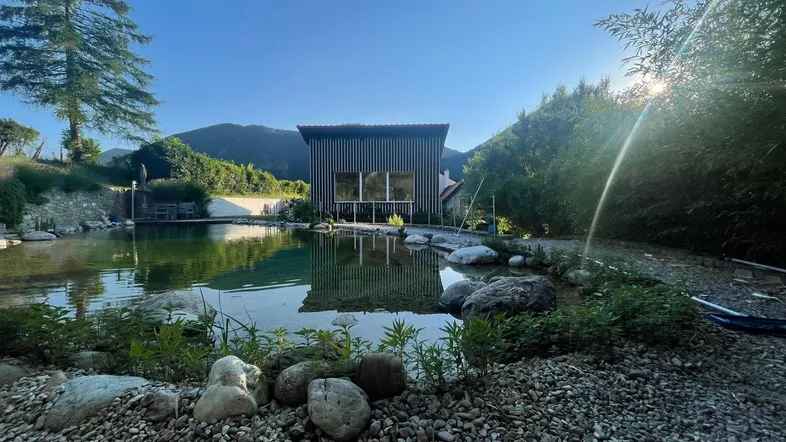 Natürlicher Schwimmteich mit modernem Gartenhaus und Bergblick unter strahlend blauem Himmel.