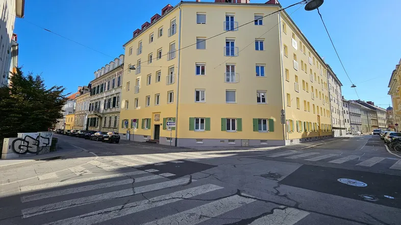 Gebäudeansicht an einer Straßenecke mit gelber Fassade und grünen Fensterläden unter blauem Himmel.