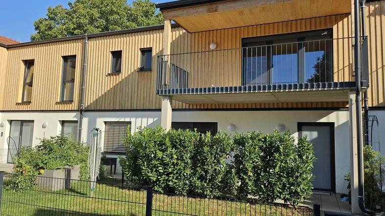 Moderne Wohnanlage mit Holzfassade, Balkonen und kleinen Gärten im Erdgeschoss unter blauem Himmel.