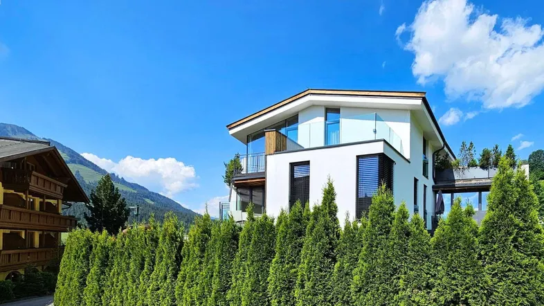 Moderne weiße Fassade eines Gebäudes mit großen Fenstern und Balkonen vor einer Bergkulisse.