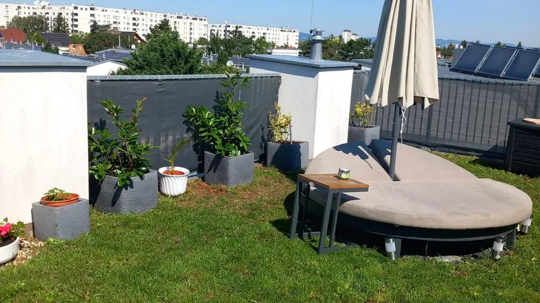 Grüne Dachterrasse mit Rasenfläche, Pflanzen in Töpfen und einem gemütlichen Tagesbett unter einem Sonnenschirm.