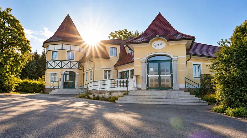 Historisches Gebäude mit markanten Türmen und rotem Ziegeldach, umgeben von viel Grün und Sonnenschein.