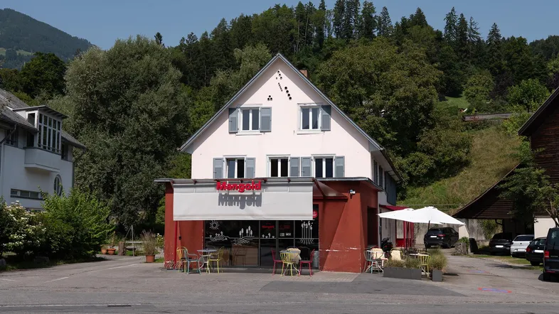 Ansicht eines Gebäudes mit Ladenfront, umgeben von Bäumen und Bergen im Hintergrund.