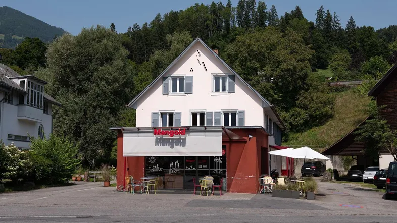 Vorderansicht eines Gebäudes mit einem Café im Erdgeschoss und Wohnungen darüber, umgeben von Bäumen.