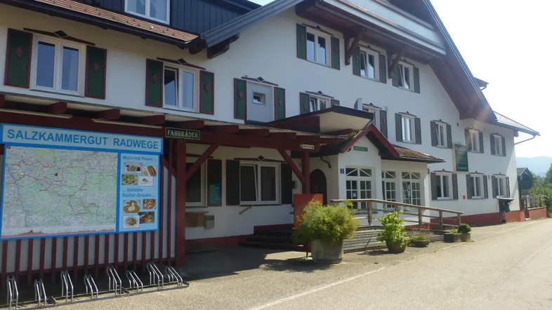 Traditionelles Gebäude mit weißer Fassade, grünen Fensterläden und einem Informationsstand für Radwege vor dem Eingang.
