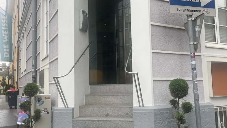 Eingangsbereich eines Gebäudes mit Steinstufen, Geländer und Straßenschildern in einer städtischen Umgebung.