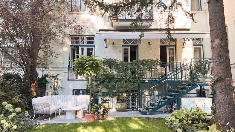 Gepflegter Gartenbereich mit Sitzgelegenheiten, Treppe zum Gebäude und üppiger Bepflanzung.