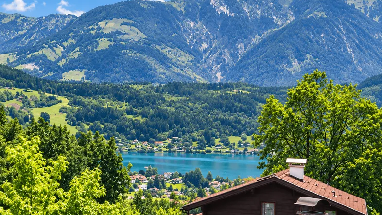 Malerischer Blick auf einen See und die umliegenden Berge mit einem traditionellen Hausdach im Vordergrund.