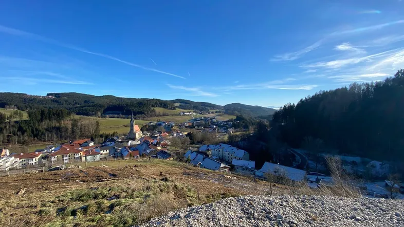 Weitläufiger Blick über ein malerisches Dorf in einem Tal, umgeben von Wäldern und Hügeln unter blauem Himmel.