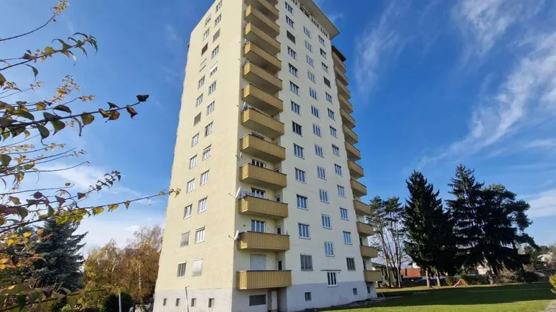 Hohes Wohngebäude mit vielen Balkonen und umliegender Grünfläche unter blauem Himmel.