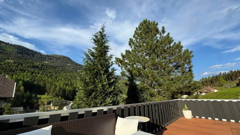 Sonnige Terrasse mit Holzboden, bequemen Sitzmöbeln und einem weiten Blick auf die grünen Berge.