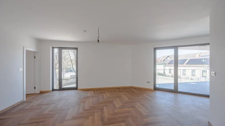 Helles, leeres Wohnzimmer mit Fischgrätparkett und großen Fenstern, die viel Tageslicht hereinlassen.