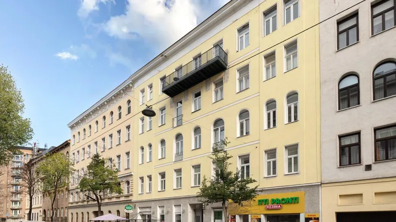 Gelbe Fassade eines Mehrfamilienhauses an einer belebten Straße mit Geschäften im Erdgeschoss.