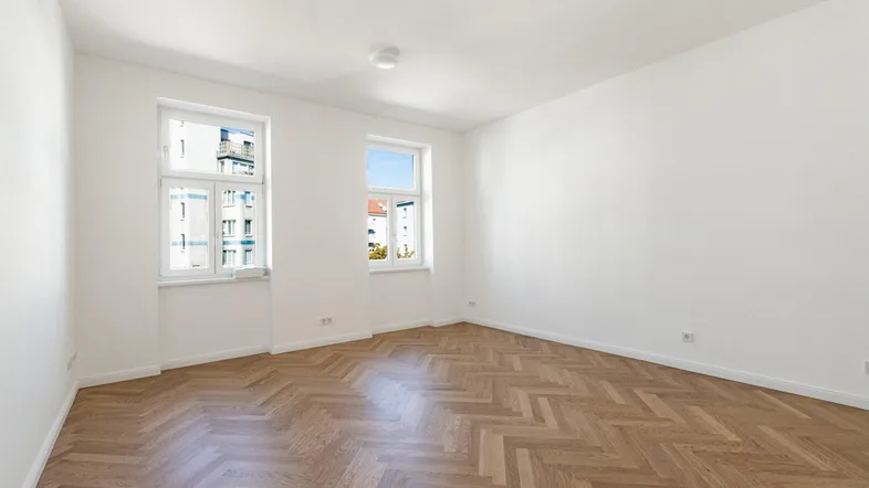 Helles, leeres Zimmer mit Fischgrätparkettboden und zwei Fenstern, die Tageslicht hereinlassen.