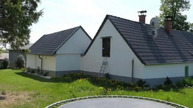 Außenansicht eines Hauses mit dunklem Ziegeldach, weißer Fassade und grünem Gartenbereich mit Trampolin.