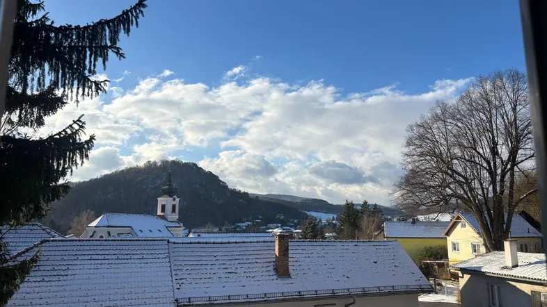 Blick aus dem Fenster auf eine verschneite Landschaft mit Kirche und Bergen unter blauem Himmel.