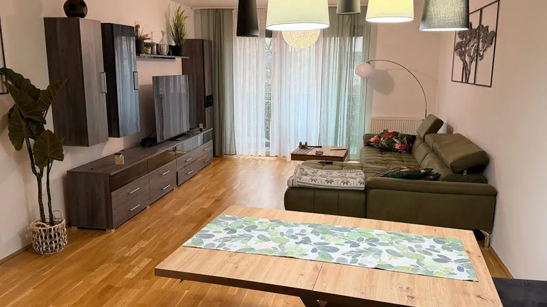 Geräumiger Wohn-Essbereich mit Parkettboden, bequemer Couch und Zugang zum Balkon durch große Fenster.