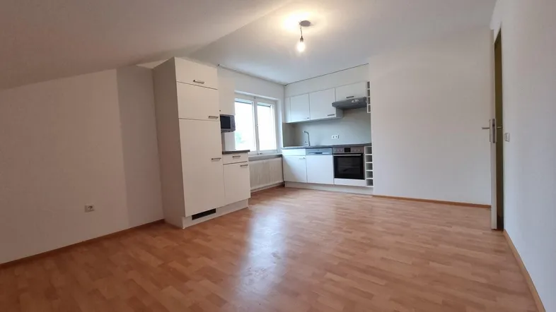 Heller offener Wohnbereich mit integrierter weißer Küche, Dachschrägen und Laminatboden.