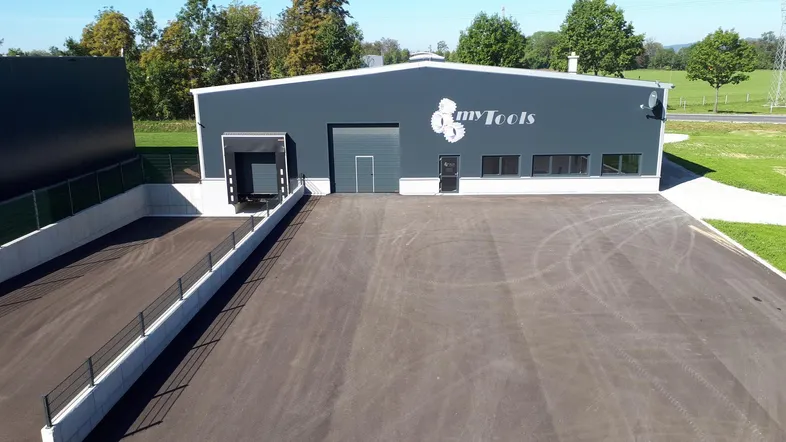 Modernes Industriegebäude mit dunkelgrauer Fassade und großem asphaltierten Vorplatz, umgeben von Grünflächen und Bäumen.