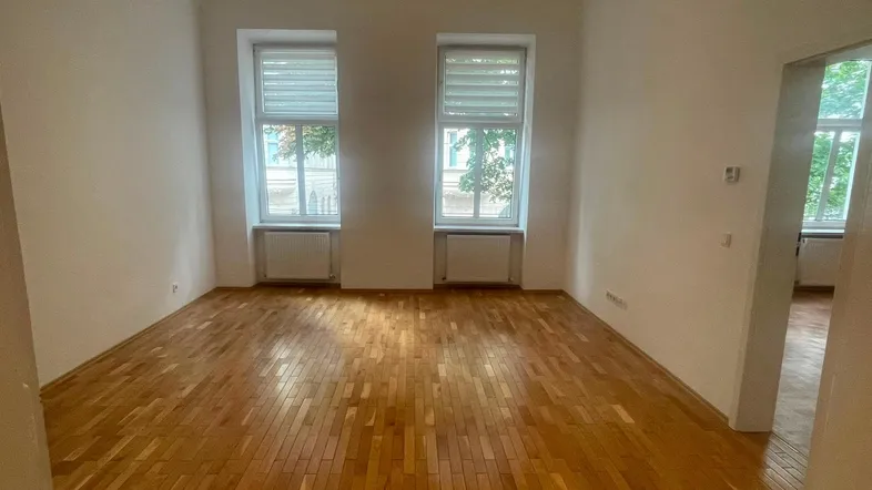 Helles, leeres Zimmer mit Parkettboden und zwei Fenstern, bereit zur individuellen Gestaltung.