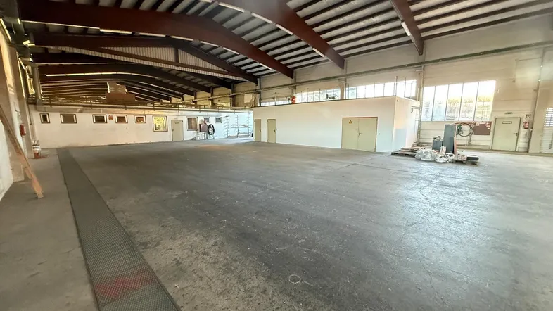 Große, leere Industriehalle mit hohen Decken, sichtbaren Holzbalken und Betonboden, gut beleuchtet durch Fenster.