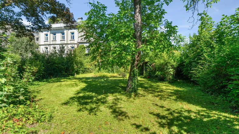 Grüner Gartenbereich mit Rasenfläche und Bäumen, im Hintergrund ist ein historisches Gebäude sichtbar.