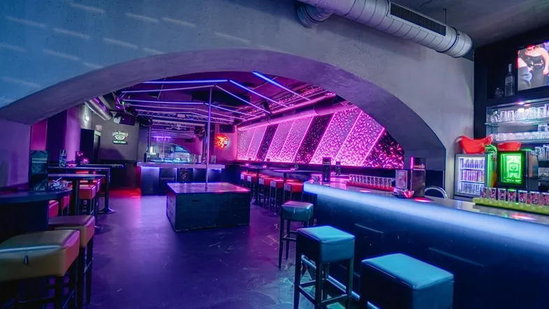 Moderne Bar mit leuchtenden Neonlichtern, Tanzstange und gemütlichen Sitzbereichen in Lila- und Blautönen.