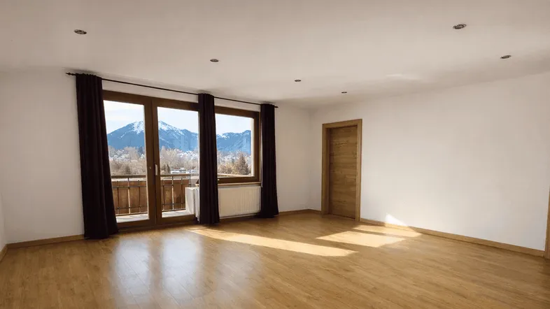 Helles, leeres Zimmer mit Holzboden und Zugang zu einem Balkon mit Bergblick.