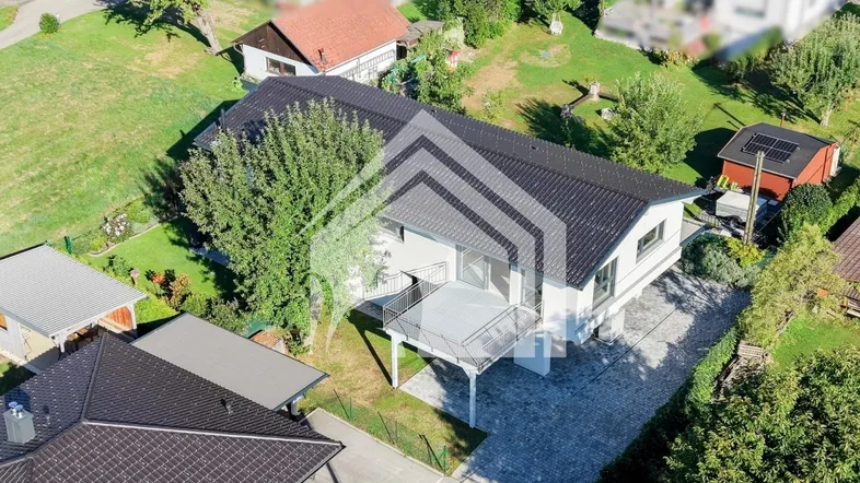 Luftaufnahme eines modernen Hauses mit dunklem Dach, weißer Fassade und großem Balkon in grüner Umgebung.