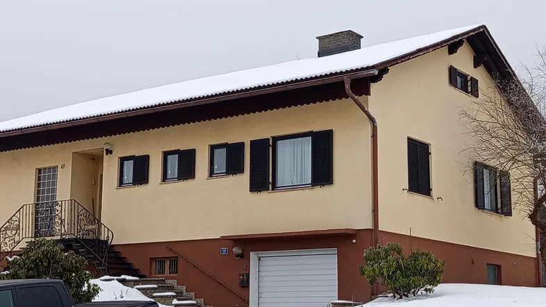Traditionelles Einfamilienhaus mit beiger Fassade, braunem Sockel und verschneitem Dach im Winter.