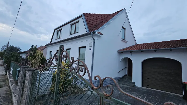 Weiß verputztes Einfamilienhaus mit rotem Ziegeldach und Garagenzufahrt unter bewölktem Himmel.